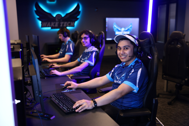 Eine Gruppe von Menschen in Headsets lächelt, während sie bei einem Esport-Turnier-Setup an Computern arbeiten, mit Monitoren, Tastaturen und einer markierten Wand im Hintergrund.