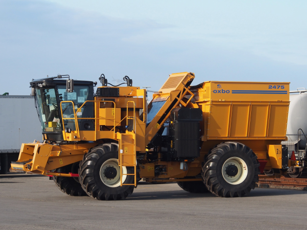 Großer gelber OXBO 2475 Radlader vor einem weißen Container auf einer Straße mit einem klaren blauen Himmel im Hintergrund geparkt.