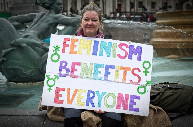 Eine Frau Setzt Sich An Einer Fontäne Hin, Holding Ein Schild Mit Der Aufschrift "Feminismus Beruht Auf Jeden" Mit Einer Tasche Neben Ihr, Während Verwischte Gestalten, Eine Statue Und Ein Gebäude Im Hintergrund Erscheinen.