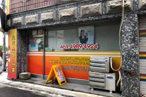 Ein Gebäude mit einer Tafel, die "Koreanische Klimaservice" anzeigt, einem Rollladen, einer Tafel mit Text und einem Personenbild, verschiedenen auf dem Boden liegenden Geräten, Pfosten, Bäumen, einem Auto auf der Straße und Drähten unter einem bewölkten Himmel.