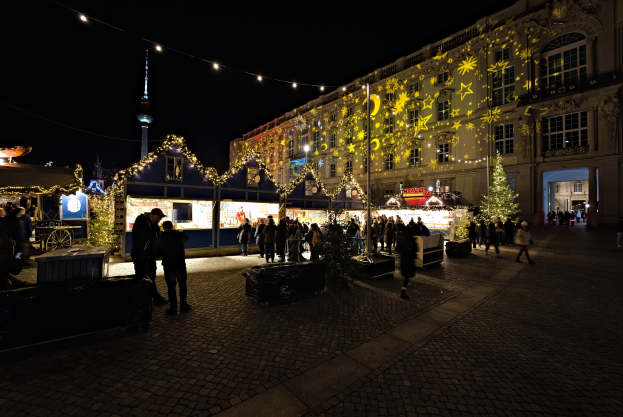 Ein geschäftiger Weihnachtsmarkt in Berlin, Deutschland mit Menschen um dekorierte Stände, festliche Lichter und dunklem Himmel mit Gebäuden im Hintergrund.