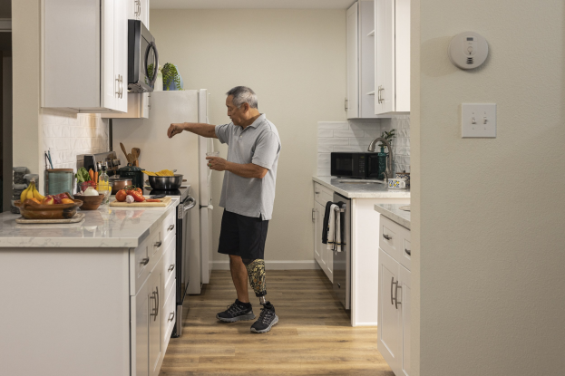 Ein Mann steht in einer Küche mit weißen Schränken und hält ein Glas in der Hand, während verschiedene Gegenstände wie Obst, Gemüse und Utensilien auf einer Arbeitsplatte zu seiner Linken liegen.