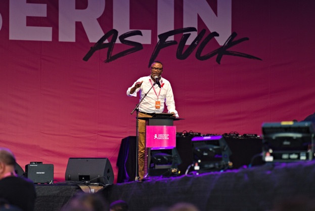 Mann in weißem Hemd und braunen Hosen, der in ein Mikrofon auf einer Bühne spricht, mit einem pinken "Berlin as Fuck"-Schild dahinter.