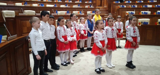 Eine Gruppe von Kindern in weißen und roten Kleidern steht zusammen in einem Raum, mit einem Podium und einem Mikrofon auf der linken Seite, Stühlen und Tischen im Hintergrund und Fahnen, die eine nationale Versammlung anzeigen.