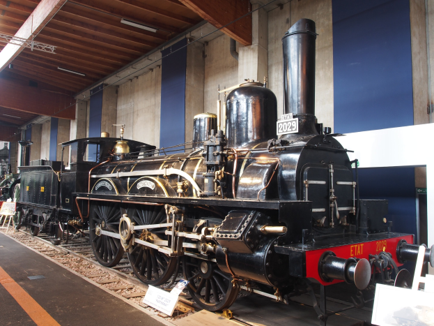 Alte Dampflokomotive in einem Museums-Setting mit umgebenen Eisenbahnschienen, Informationsschildern und anderen Ausstellungsstücken, vor einer Wand mit Deckenbeleuchtung.