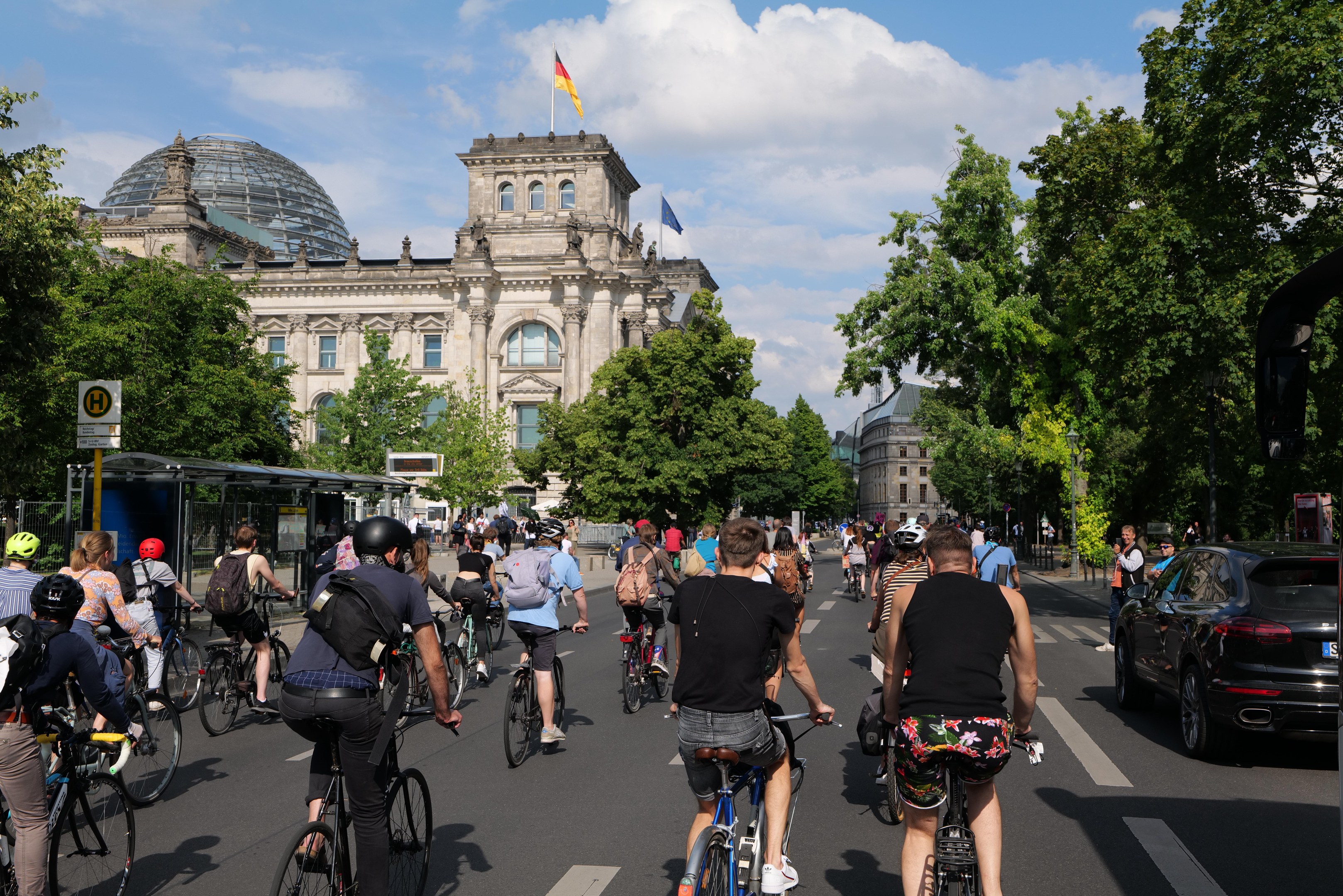 Eine Gruppe von Menschen, die auf Fahrrädern eine von Bäumen gesäumte Straße in Berlin entlangfahren, mit Gebäuden und einer Bushaltestelle, unter einem bewölkten Himmel und einer Flagge auf einem Gebäude.