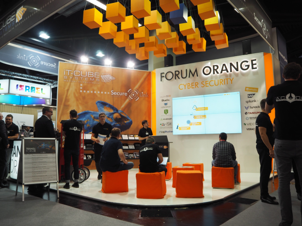 Eine Gruppe von Menschen an einem IT-Cube Systems Forum Orange Cyber Security Stand auf einer Messe, mit Stühlen, stehenden Besuchern, einer Tafel, einem Display, Regalen, Schildern, Deckenleuchten und Lampen.