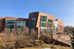Großes mehrstöckiges Gebäude mit zahlreichen Fenstern, umgeben von Bäumen, Bänken, Gras und einem Laternenpfahl, mit anderen Gebäuden und einem klaren blauen Himmel im Hintergrund, als der neue Campus der University of Illinois at Chicago identifiziert.