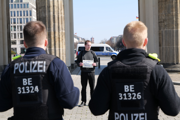 Drei Polizeibeamte vor einem Gebäude mit einem Mann, der ein Blatt Papier zwischen ihnen hält, flankiert von Säulen, mit Fahrzeugen, Menschen, Bäumen, Gebäuden und Himmel im Hintergrund.