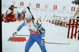 Eine Frau in einer blauen und weißen Kleidung fährt einen schneebedeckten Hügel mit Skiern hinunter und hält Ski-Stöcke in der Hand, im Hintergrund sind Menschen, eine Kamera auf einem Stativ und eine Nummern-Tafel zu sehen.