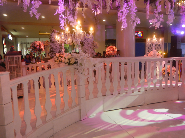 Weiße Reling mit Blumen und Kronleuchtern hängt von der Decke bei einer Hochzeitsfeier, mit Menschen drumherum, Säulen und Lichtern im Hintergrund und einem Bildschirm auf der rechten Seite.