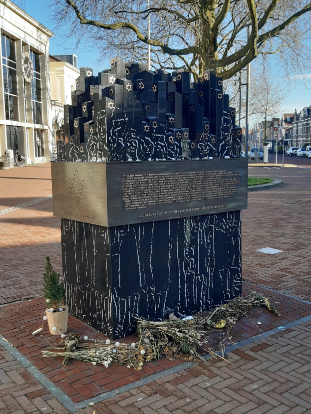 Denkmal in einem Stadtplatz für Holocaust-Opfer mit einem Baum davor, einem Topfgewächs auf dem Boden, verstreuten trockenen Blättern und Gebäuden, Fahrzeugen und Bäumen im Hintergrund.