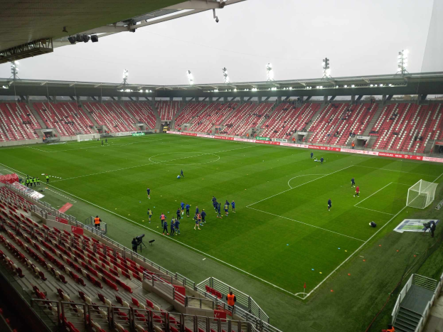Ein Fußballfeld in einem Stadion mit Menschen, Sitzplätzen, Geländern, Beleuchtung und einem Himmel als Hintergrund.