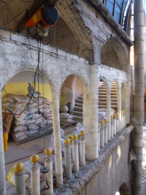 Altes Gebäude mit Bogengang, Säulen und Treppen wird renoviert, mit verstreuten Werkzeugen, Materialien und Gegenständen, darunter Taschen und eine Maschine mit Drähten oben drauf.