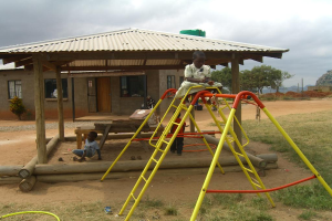 Ein Mädchen sitzt auf einer Eisenstange vor einem Haus mit einem Fenster und einer Tür, während ein Junge im Sand spielt.