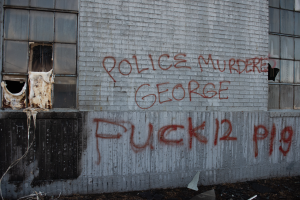 Ein Gebäude mit der Graffiti-Beschriftung "Polizei Mord George Fuck 12 P19" an der Seite, das Fenster und verschiedene Gegenstände vorne zeigt.