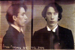 Ein Mugshot eines Mannes in einem schwarzen Anzug, weißem Hemd und schwarzer Krawatte mit ernstem Gesichtsausdruck und zurückgekämmten Haaren, begleitet von zwei weiteren Fotos und fetter Schrift, die seine Verbrechen beschreibt.