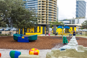 Ein städtischer Spielplatz mit Kindern auf der Ausrüstung, umgeben von Bäumen und hohen Gebäuden, mit Fahrzeugen auf der Straße und Himmel darüber.