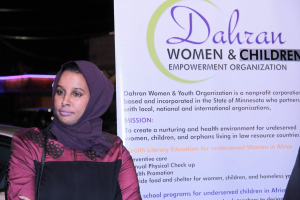 Eine Frau in einem weißen Hemd und blauen Jeans steht entschlossen vor einer Tafel, auf der "Dahran Frauen & Kinderermächtigungsorganisation" steht, mit einem Auto im Hintergrund.