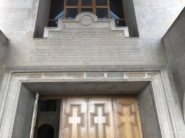 Große hölzerne Tür vor einem steinernen Gebäude mit einem Glasfenster darüber, das likely eine Inschrift trägt, die Holocaust-Opfer gedenkt.