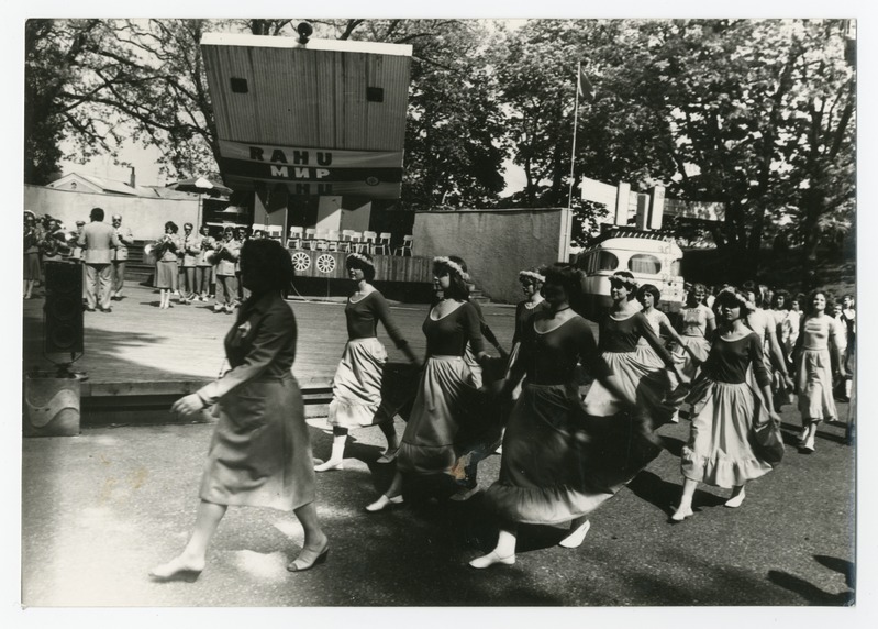 Eine Gruppe von Frauen, die auf der Straße tanzen, umgeben von einer Menge, einem Lautsprecher, einem Fahrzeug, einer Tafel mit Text und einer Fahnenstange.