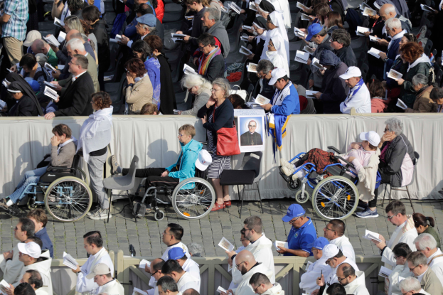 Eine große Gruppe von Menschen in Rollstühlen und Stühlen sitzt vor einer Menge, einige tragen Mützen und halten Bücher, mit einem Banner im Hintergrund, das ein Personenfoto und Geländer zeigt.