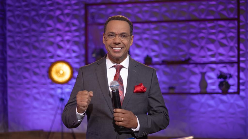 Ein Mann in Anzug und Krawatte, identifiziert als Stephen Colbert, steht auf einer Bühne und hält ein Mikrofon, während er eine Brille trägt und ein Wandständer und eine Lampe hinter ihm zu sehen sind.