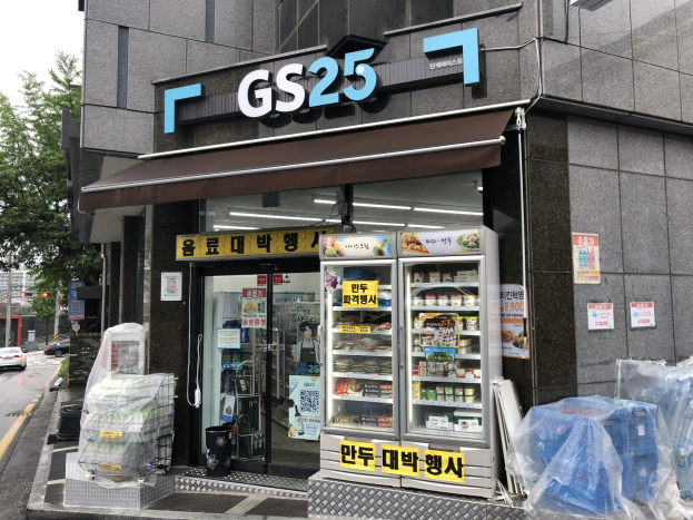 Ein Gebäude mit einer "GS25"-Aufschrift, umgeben von Fahrzeugen, Bäumen und einem klaren blauen Himmel, das einen koreanischen Lebensmittelmarkt mit Kühlgeräten beherbergt, die mit Lebensmitteln gefüllt sind.