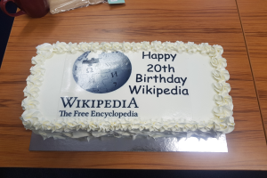 Ein Geburtstagskuchen mit "Herzlichen Glückwunsch zum 20. Geburtstag Wikipedia" darauf, der auf einem Tisch mit einer Tasse, einer Hülle und anderen Gegenständen steht.
