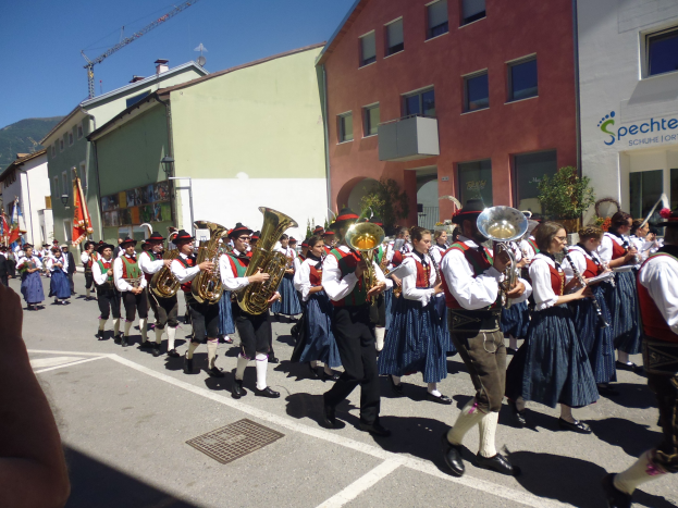 Eine Gruppe von Menschen in traditioneller bayrischer Tracht, die Musikinstrumente spielen, während sie eine Straße mit Gebäuden entlanggehen, einige halten Fahnen, mit einem Hügel und einem blauen Himmel im Hintergrund.