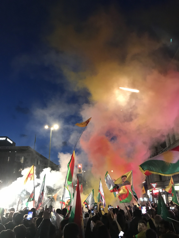 Menschenmenge vor einem Gebäude mit Fahnen und Handys, während bunte Feuerwerke den Himmel erleuchten.