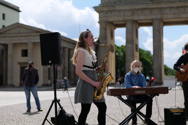 Eine Frau spielt Saxophon vor dem Reichstagsgebäude in Berlin, Deutschland, mit einer Person, die ein Piano spielt, einer anderen, die eine Gitarre hält, und einem Lautsprecher im Vordergrund, während Gebäude, Bäume und Wolken den Hintergrund füllen.