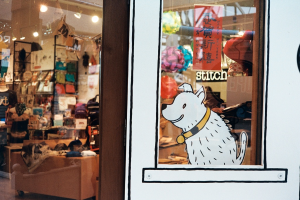 Eine Illustration eines Hundes mit Text auf der rechten Seite und auf der linken Seite ein Raum mit Lampen, Flaggen, Regalen, Sofas, Kleidung, Deko, Menschen, Stühlen und verschiedenen anderen Gegenständen.