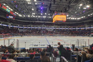 Eine große Menschenmenge in einer Hockeyarena, mit einigen sitzend und anderen stehend, die ein Spiel unter hellen Scheinwerfern und Bannern beobachten.