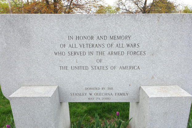 Gedenkstein mit Inschrift, die US-Militärveteranen ehrt, umgeben von Pflanzen, Blumen, Gras, Bäumen und einem bewölkten Himmel.