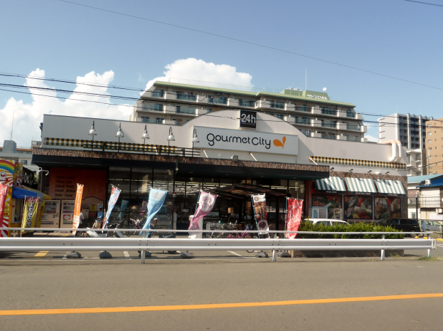 Eine belebte Stadtstraße mit Gebäuden, Straßenmöbeln, Fahrzeugen, Grünflächen und einem Restaurant, das Speisen in seinem Schaufenster präsentiert.