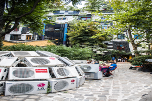 Luftkühler am Boden links, ein Weg rechts daneben, Menschen auf Stühlen neben dem Weg sitzen, mit Gebäuden, Bäumen und Pflanzen oben.