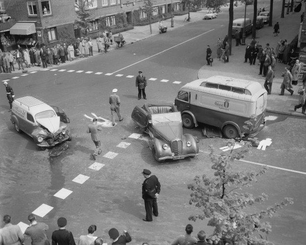 Eine Schwarz-Weiß-Szene eines Autounfalls an einer Straßenecke, mit mehreren Fahrzeugen, darunter einem Lieferwagen, einer Gruppe von Menschen drumherum und im Hintergrund Gebäude, Bäume, Masten und Texttafeln.